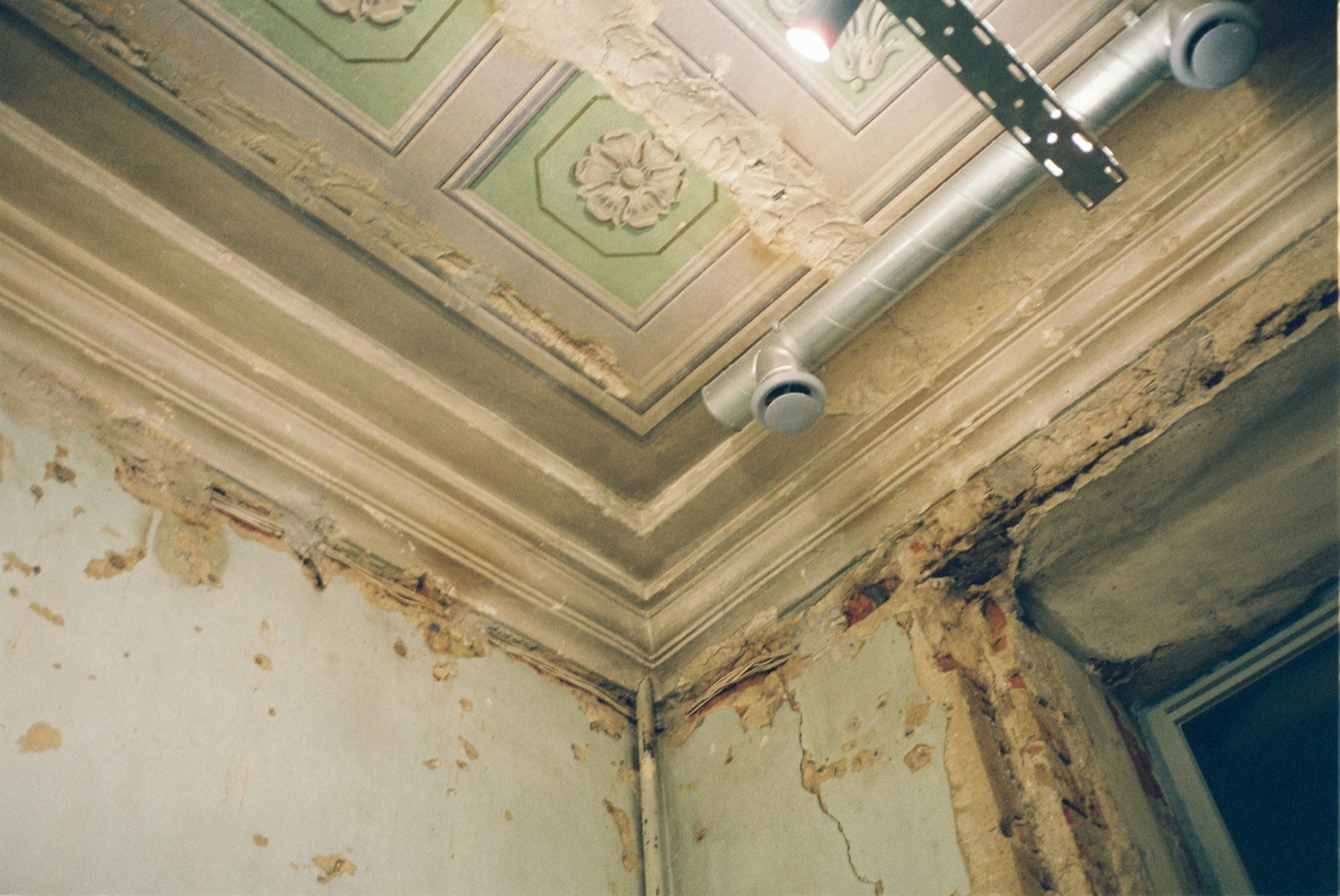 A water stained ceiling and wall corner with plaster peeling away and exposed ductwork, showing long running moisture damage