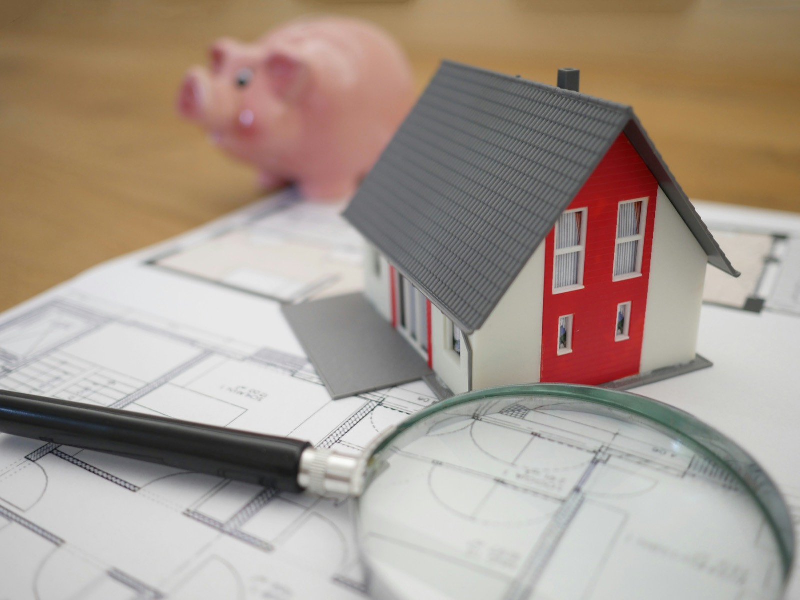 A model house and a magnifying glass resting on a set of building plans, with a piggy bank in the background