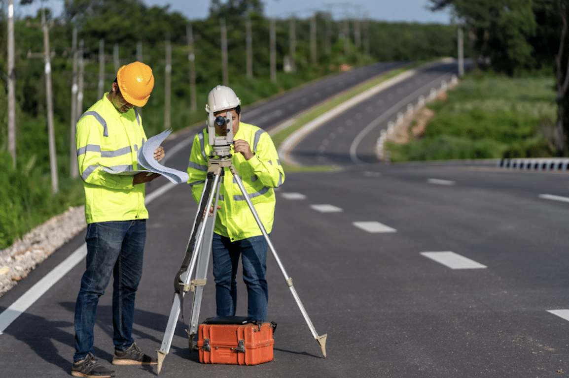 Two surveyors in high-visibility jackets using a theodolite to survey a new road