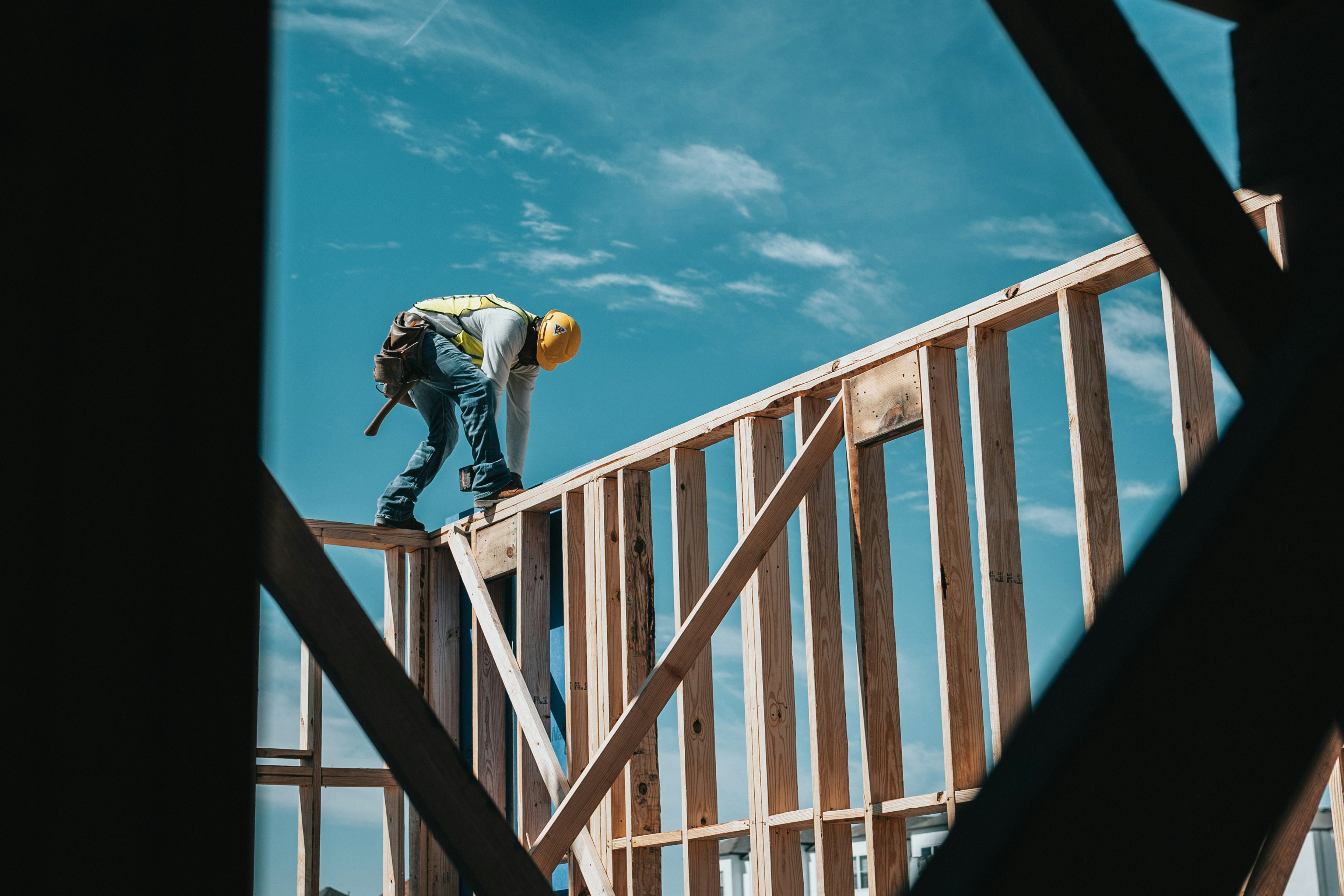 A construction worker in a hard hat and safety vest standing on the wood-framed wall of a building against a bright blue sky