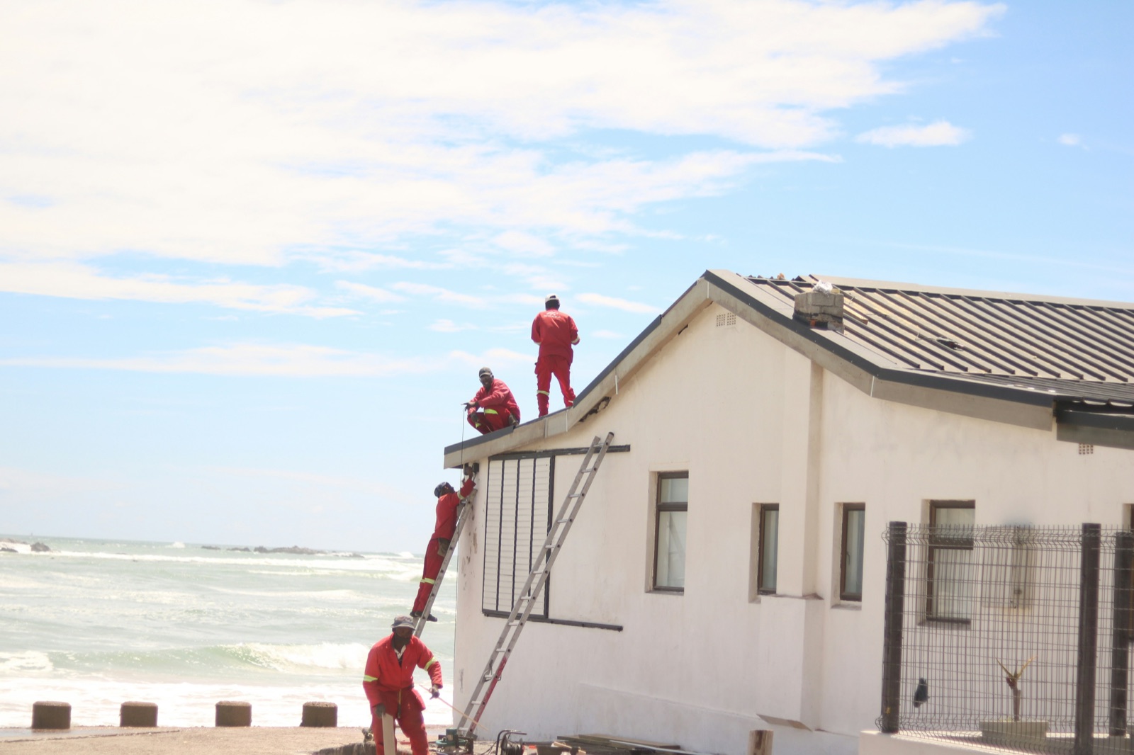 A crew in red coveralls working on the roof of a building, one on a ladder and two on the roofline, with the ocean behind them